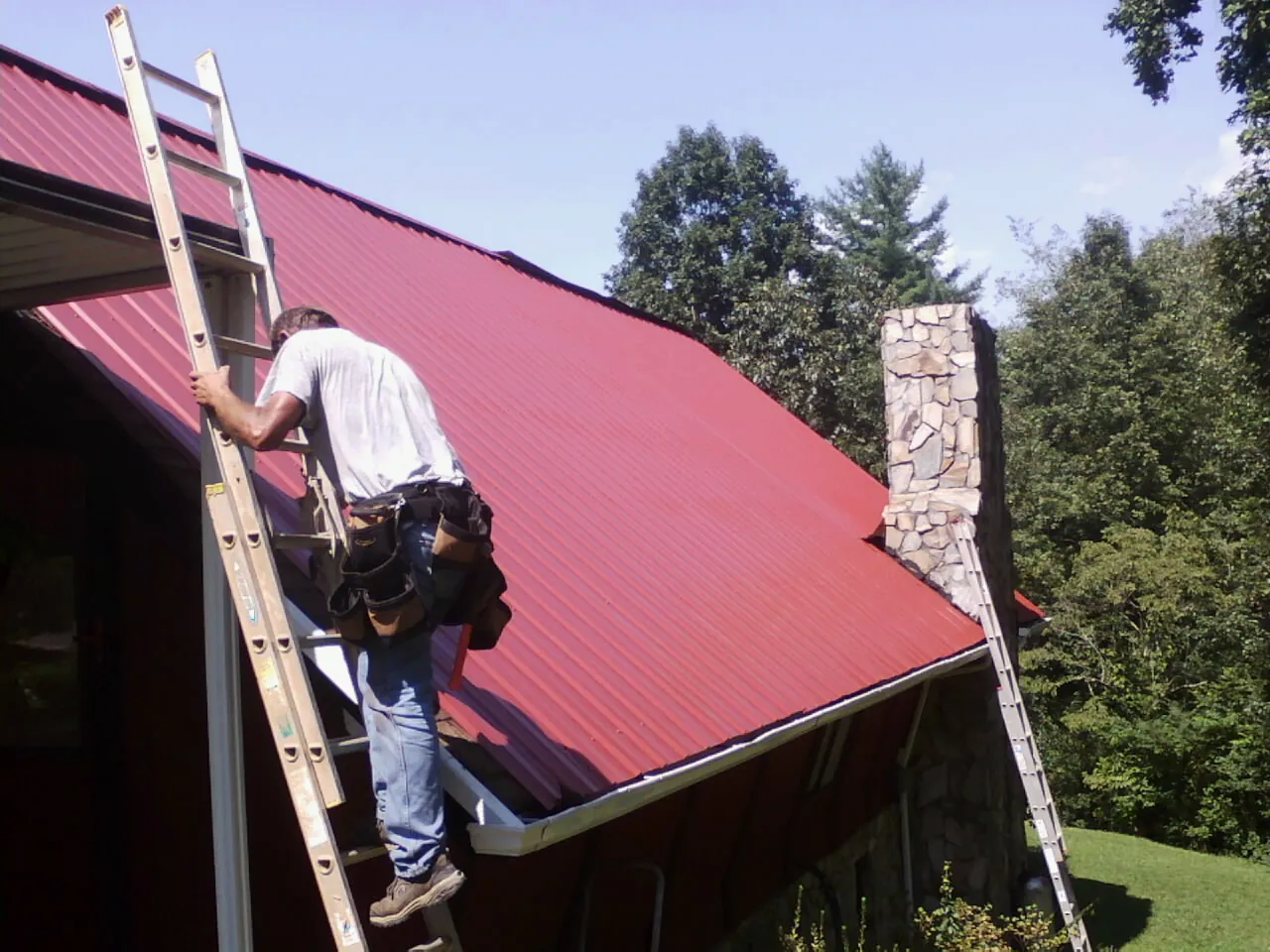 Roofer on a ladder beside a red metal roof and stone chimney — Soffit & Fascia Repair by Roofing Brothers in Bloomsburg